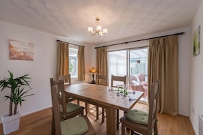 A dining area with a wooden table, chairs, a chandelier, and a view through large windows.