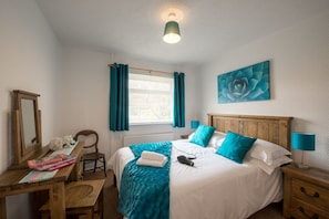 A hotel room with a bed, a desk, a chair, a window with curtains, a painting on the wall, and a lamp.