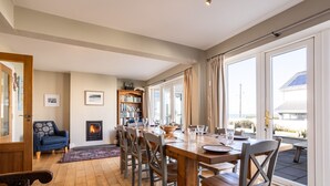 Ein Essbereich mit Holztisch, Stühlen und Kamin. Ein Bücherregal und ein Ausblick durch große Fenster sind ebenfalls vorhanden.