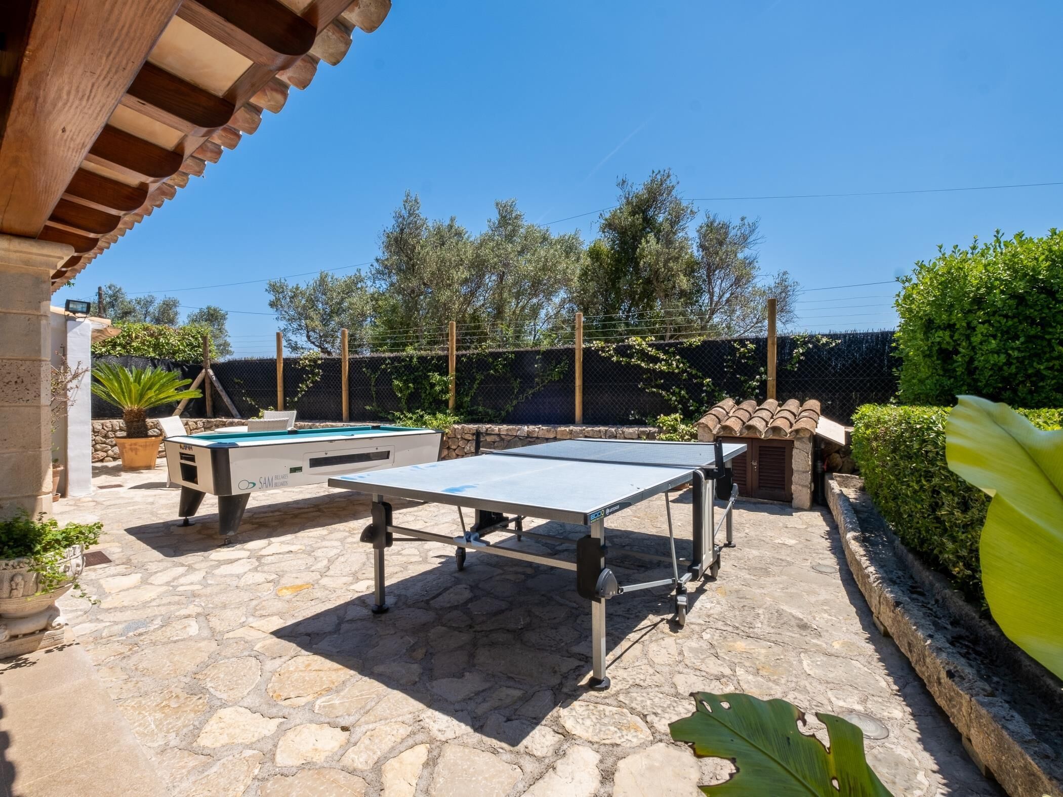 Une table de billard et une table de ping-pong se trouvent dans un espace extérieur dallé de pierre, avec une piscine et de la verdure en arrière-plan.