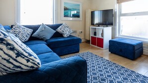 A modern living room with a blue sofa, patterned cushions, a blue ottoman, a flat-screen TV, and a framed picture on the wall. - Bryn Awel: Family-Friendly 3-Bed Apartment in Rhosneigr | Managed by Anglesey Holiday Lettings (Rhosneigr)