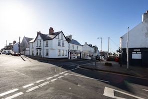 A street view with a row of white buildings, a visible storefront, and a clear road with marked lanes. - Bryn Awel: A three bed flat in Rhosneigr with sea views (Rhosneigr)