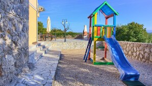 Un terrain de jeux avec un toboggan coloré et une structure d’escalade, le tout adossé à un mur de pierre et se découpant sur un ciel dégagé.