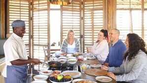 Un gruppo di persone cena in un ambiente rustico con pareti in legno, un grande tavolo apparecchiato con diversi piatti e uno chef che prepara una pietanza.