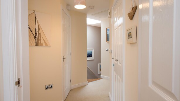 A hallway with white walls, a carpeted floor, and a ceiling light. There are doors on both sides, one with a picture frame and another with a small shelf. - Bryns Eglwys: Pet-friendly two bed holiday apartment in Rhosneigr (Rhosneigr)