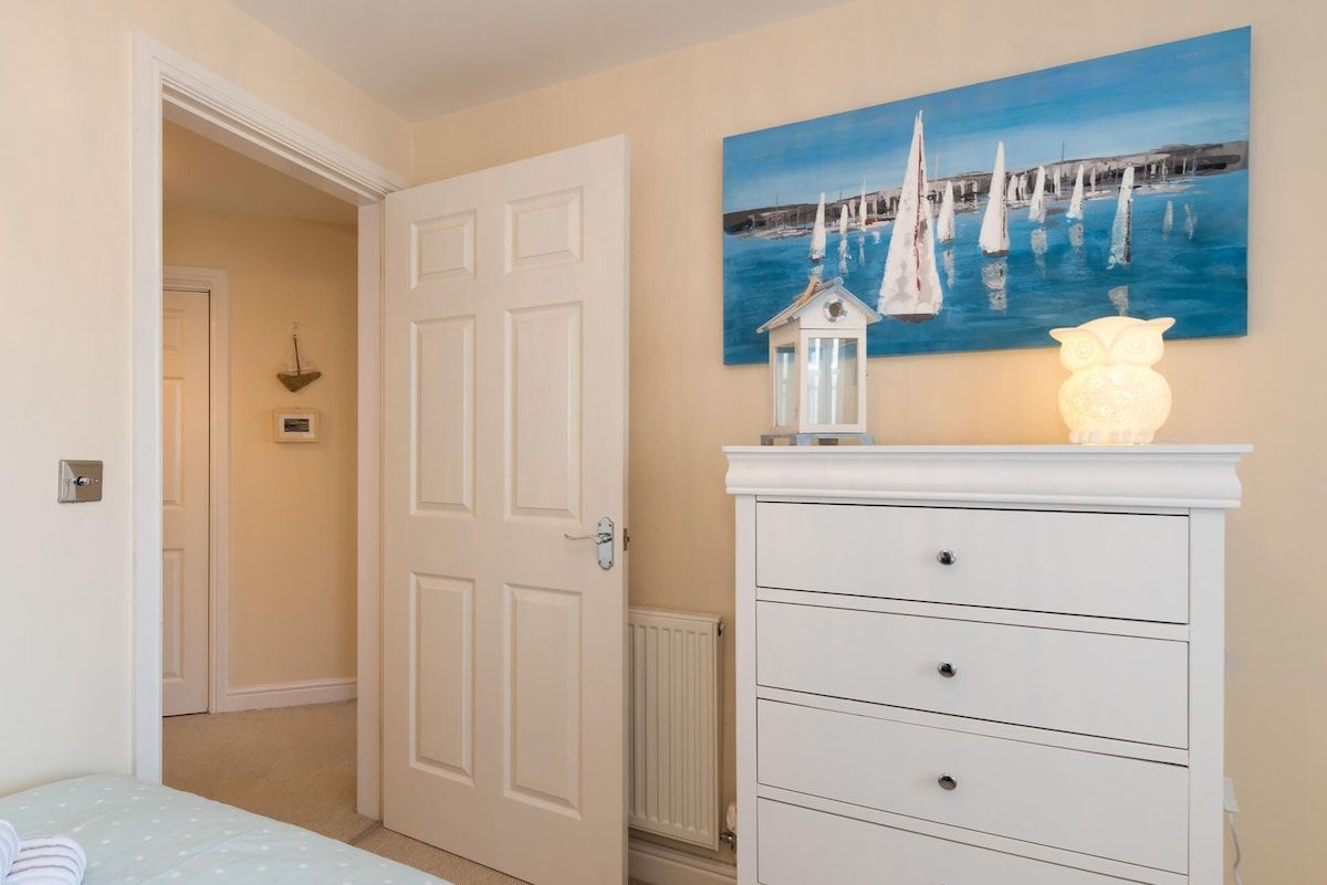 A bedroom with a bed, a white dresser, a painting of sailboats, and a lit decorative owl.