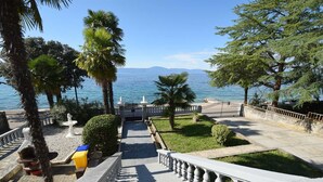 Une promenade en bord de mer agrémentée d’une balustrade en pierre, de palmiers et offrant une vue sur la mer et les montagnes lointaines.