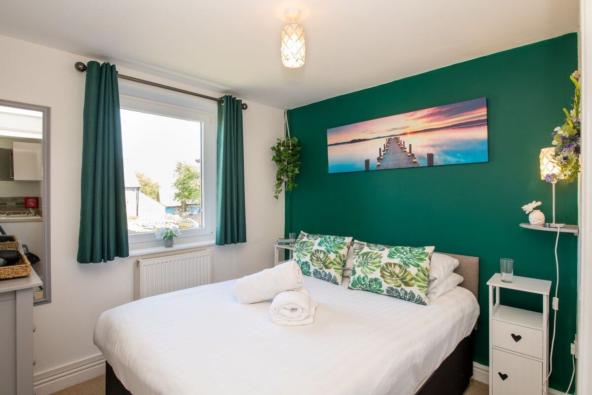 A bedroom with a bed, a window with curtains, a wall-mounted painting, and a bedside table with a lamp.