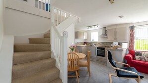 A staircase leading to a dining area with a table and chairs, a red sofa, and a kitchen in the background. - Bwthyn Siwan: Charming Pet-Friendly Cottage in Aberffraw | Managed by Anglesey Holiday Lettings (Ty Croes)