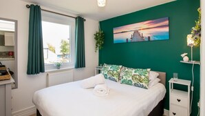 A bedroom with a bed, a window with curtains, a nightstand, a wall-mounted lamp, and a painting. - Bwthyn Siwan: Charming Pet-Friendly Cottage in Aberffraw | Managed by Anglesey Holiday Lettings (Ty Croes)