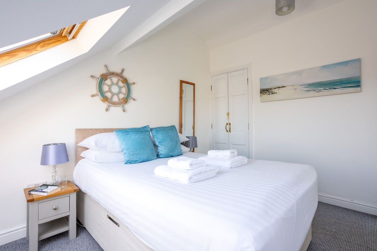 A neatly made bed with white linens and blue pillows, a small bedside table with a lamp, a ship's wheel decoration on the wall, and a beach scene painting.