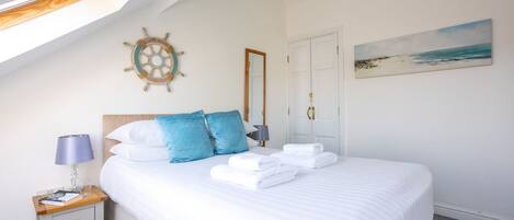 A neatly made bed with white linens and blue pillows, a small bedside table with a lamp, a ship's wheel decoration on the wall, and a beach scene painting.