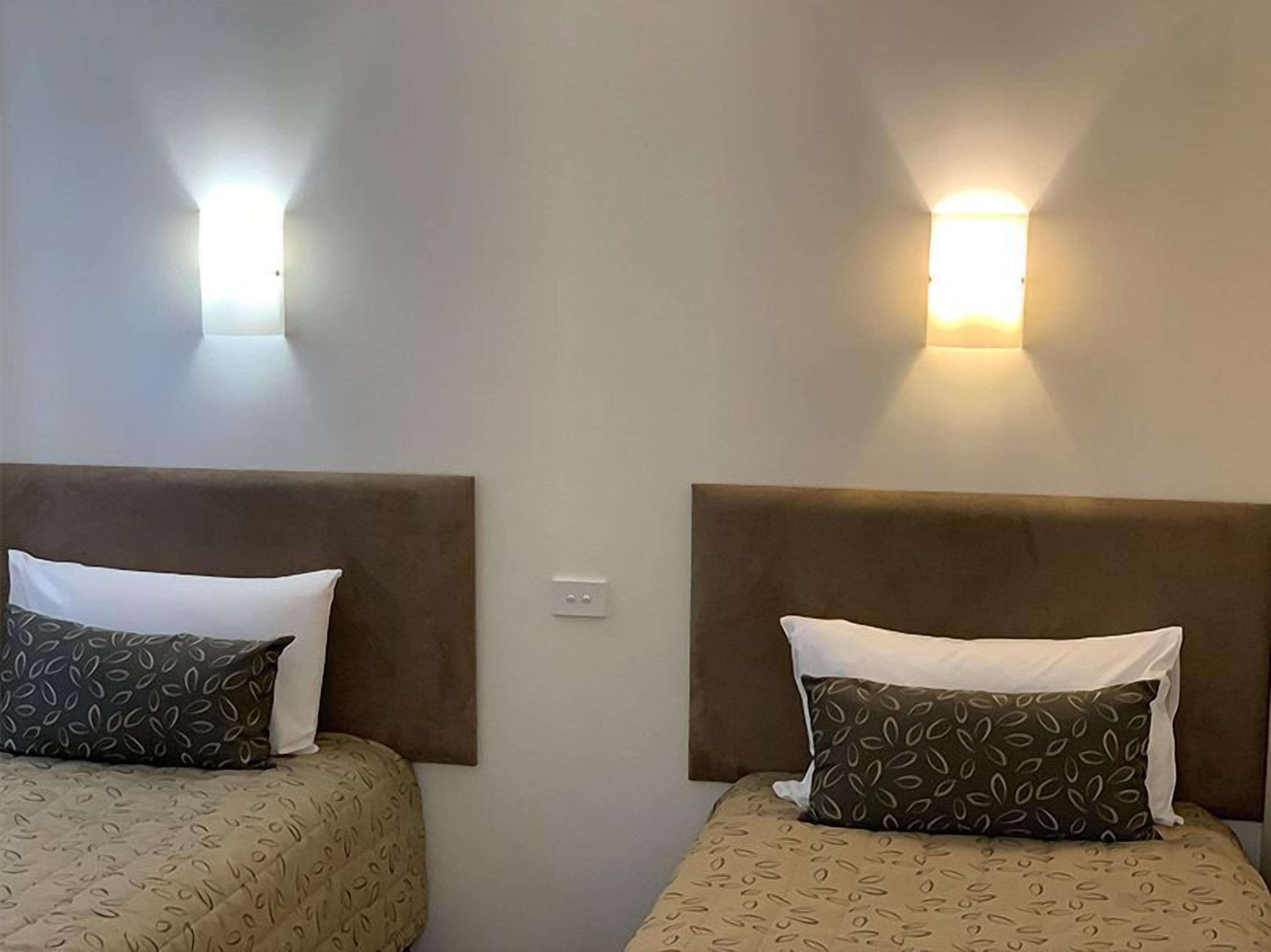 Two beds with headboards, each with a pillow and a wall-mounted light fixture.