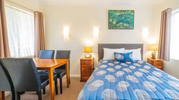 A hotel room with a bed, a desk, two chairs, a nightstand with lamps, and a painting on the wall.