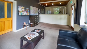 Una zona de recepción con un sofá oscuro, una puerta de madera, un tablón de anuncios con varios papeles, una mesita con revistas y una cocina al fondo.