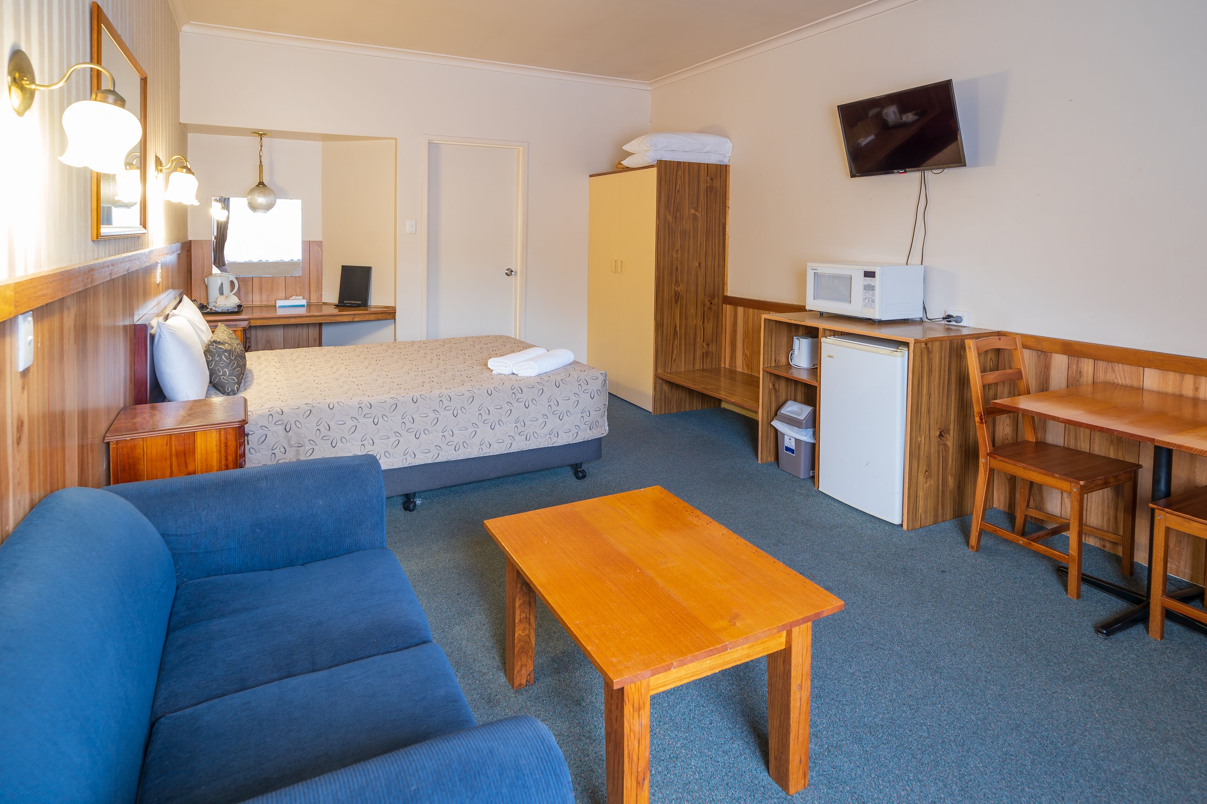 A compact hotel room with a bed, a sofa, a wooden table, a desk with a chair, a microwave, a refrigerator, and a TV mounted on the wall.