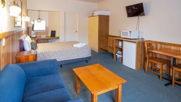 A compact hotel room with a bed, a sofa, a wooden table, a desk with a chair, a microwave, a refrigerator, and a TV mounted on the wall.