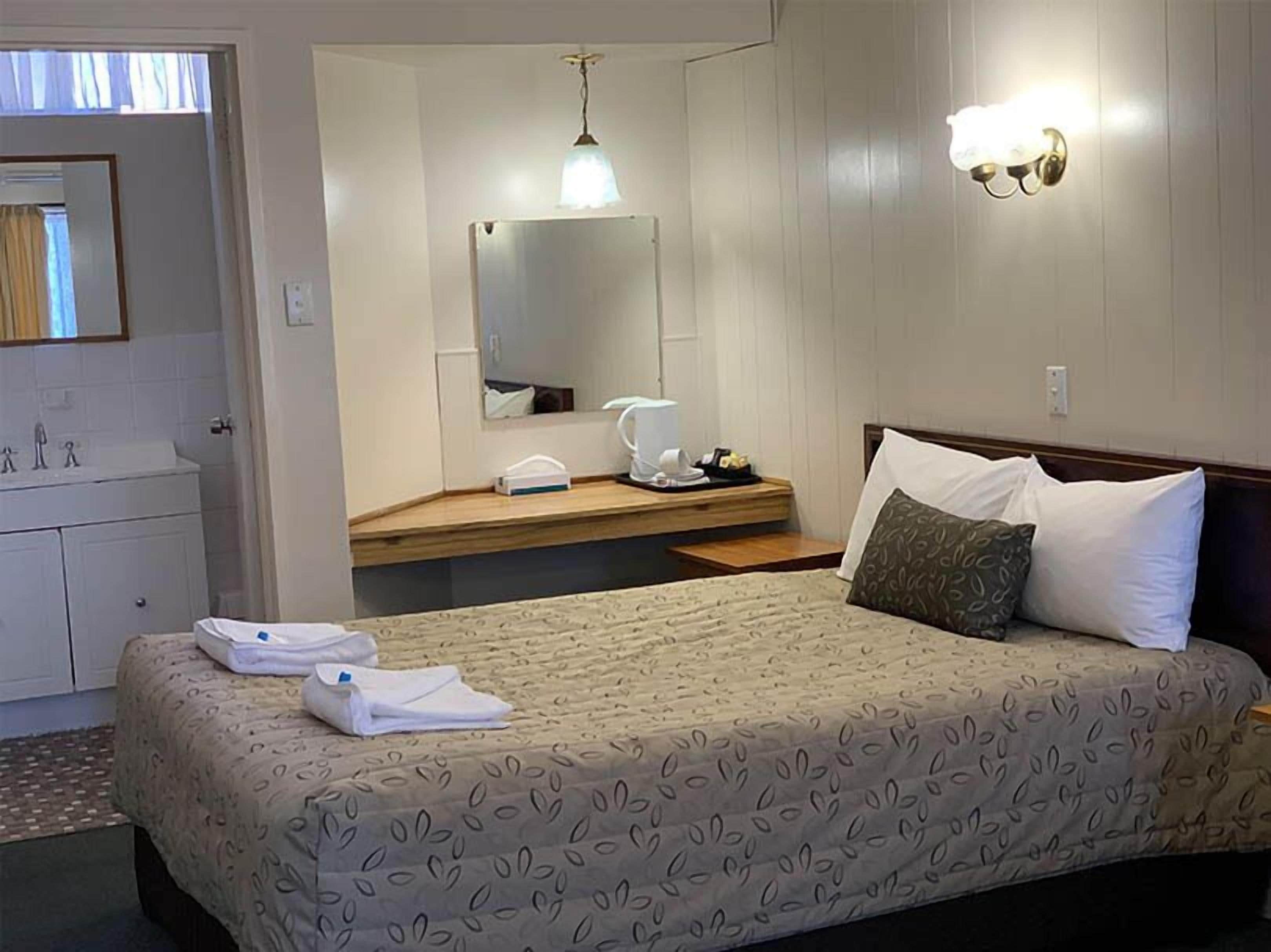 A hotel room with a bed, a bedside table, a mirror, a lamp, and a bathroom visible through an open door.