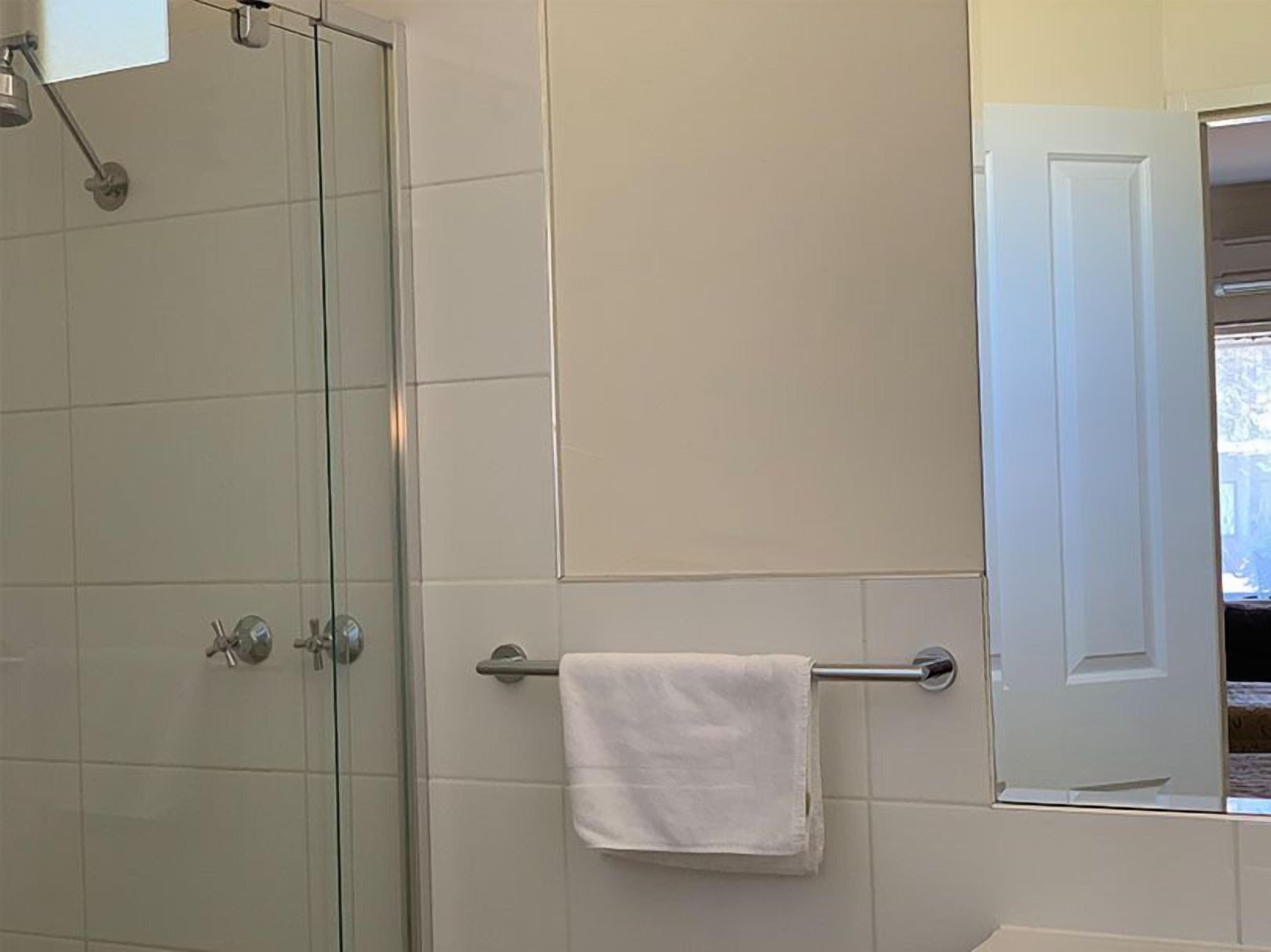 Un baño con cabina de ducha de vidrio, un perchero con una toalla blanca y una puerta que da a un pasillo.