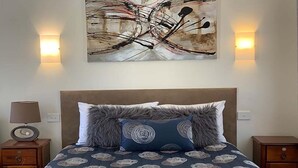 Una cama bien hecha con una colcha estampada, dos almohadas y toallas dobladas. Hay una lámpara de noche a la izquierda y una luz empotrada en la pared a la derecha.