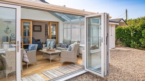 Eine offene Glasdoor führt in einen möblierten Wohnbereich mit Korbmöbeln, einem Couchtisch mit Brot und Wein und Blick auf einen Garten mit einer Hecke.