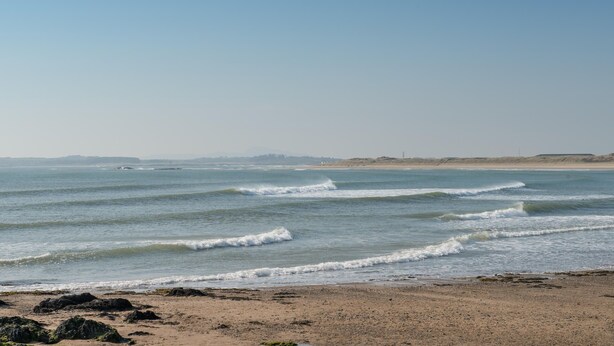 Ein Strand mit Wellen, blauem Himmel und weiter Küste.