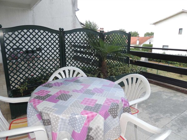 A small outdoor patio with a round table covered by a patchwork tablecloth, surrounded by white plastic chairs.