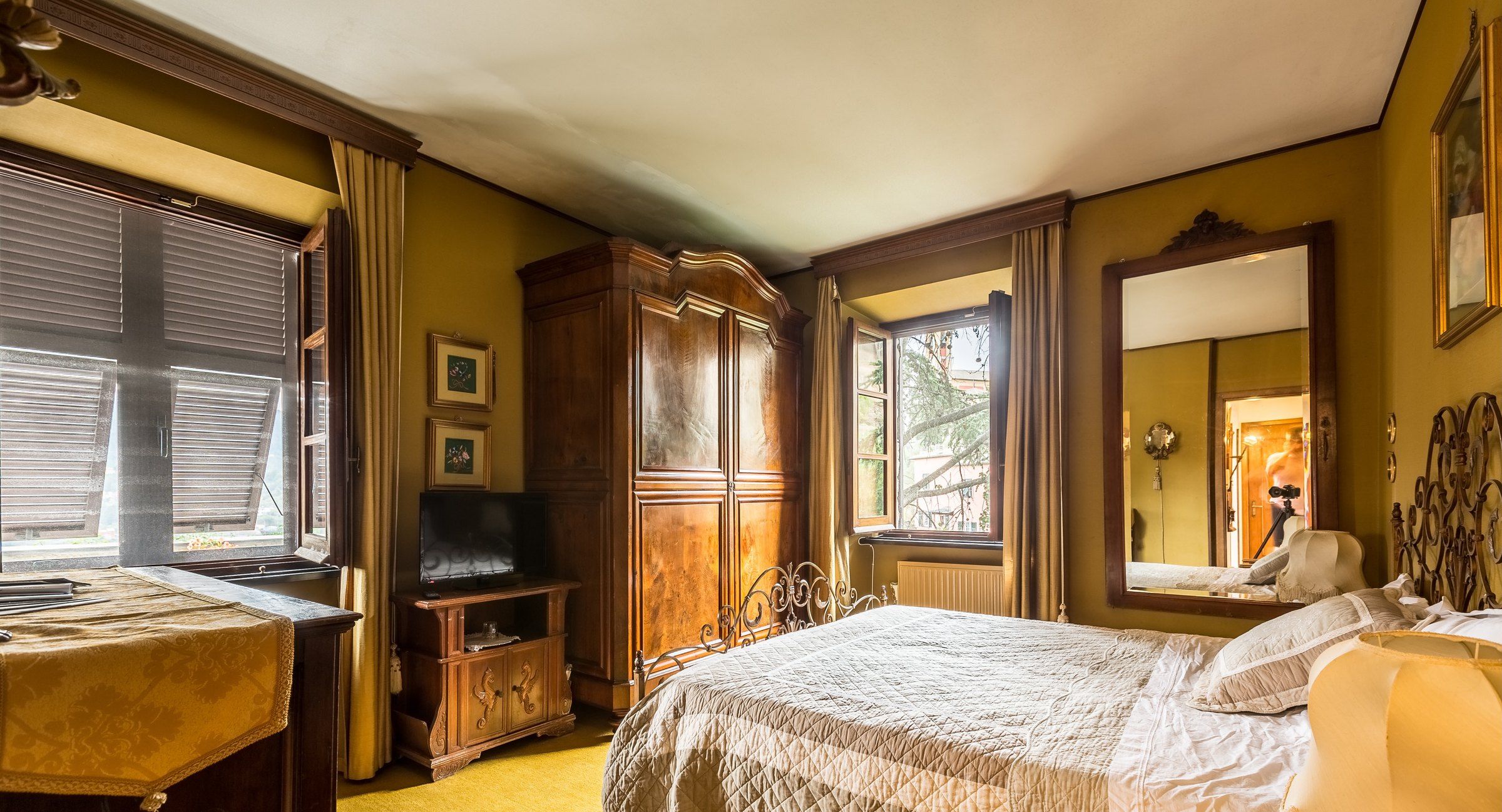 A bedroom with a bed, a wooden wardrobe, a desk with a chair, a television, and a large mirror.