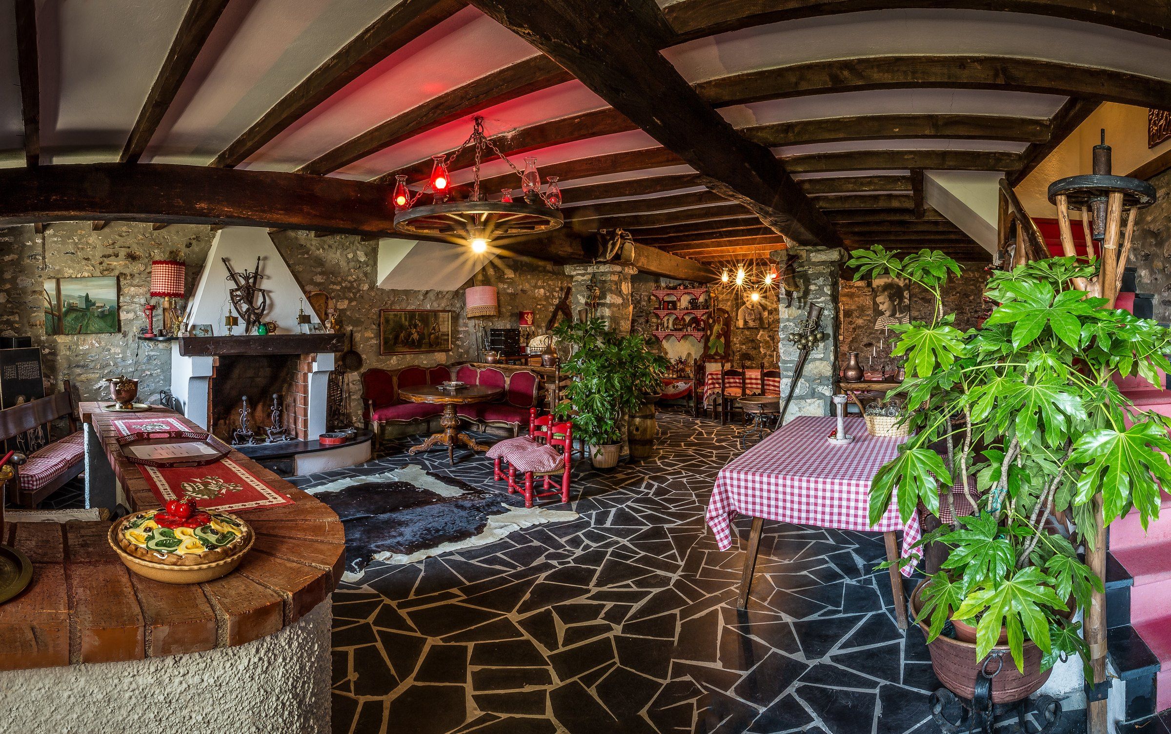 Un intérieur rustique avec des poutres apparentes, une cheminée en pierre et un coin repas agrémenté d’une nappe à carreaux.