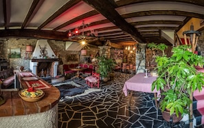 Un intérieur rustique avec des poutres apparentes, une cheminée en pierre et un coin repas agrémenté d’une nappe à carreaux.