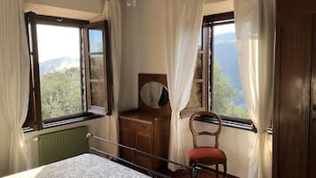 Un dormitorio con cama, cómoda de madera con espejo, una silla y dos ventanas con cortinas.