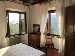 Quarto com cama, cómoda de madeira com espelho, cadeira e duas janelas com cortinas.