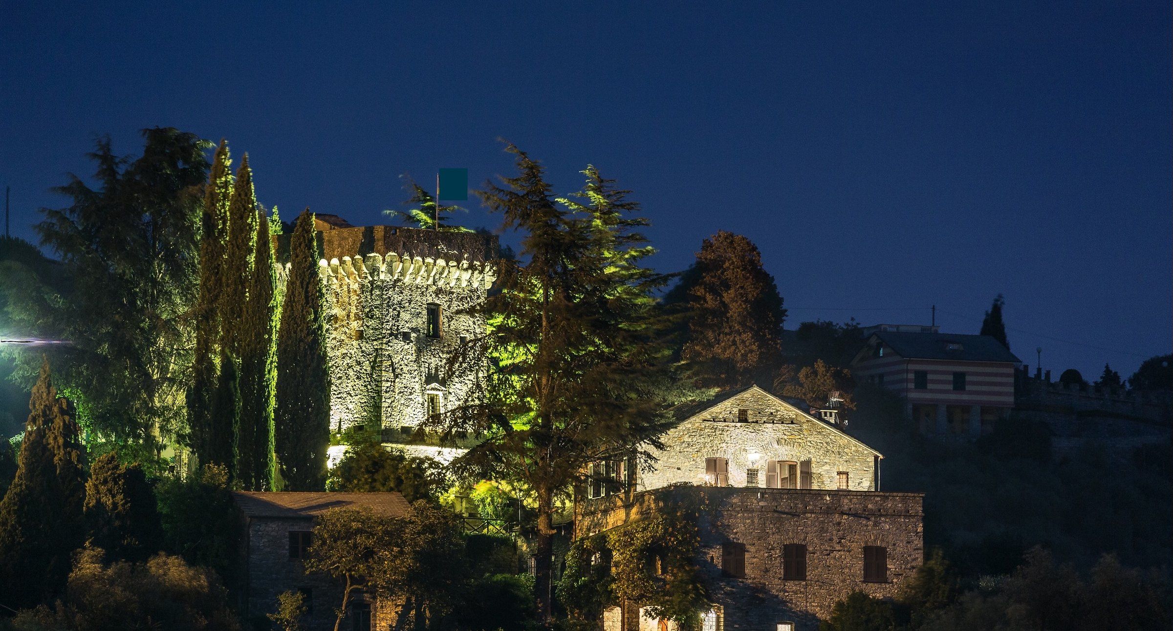 A historic stone building with a tower, surrounded by trees and a smaller building in the foreground.