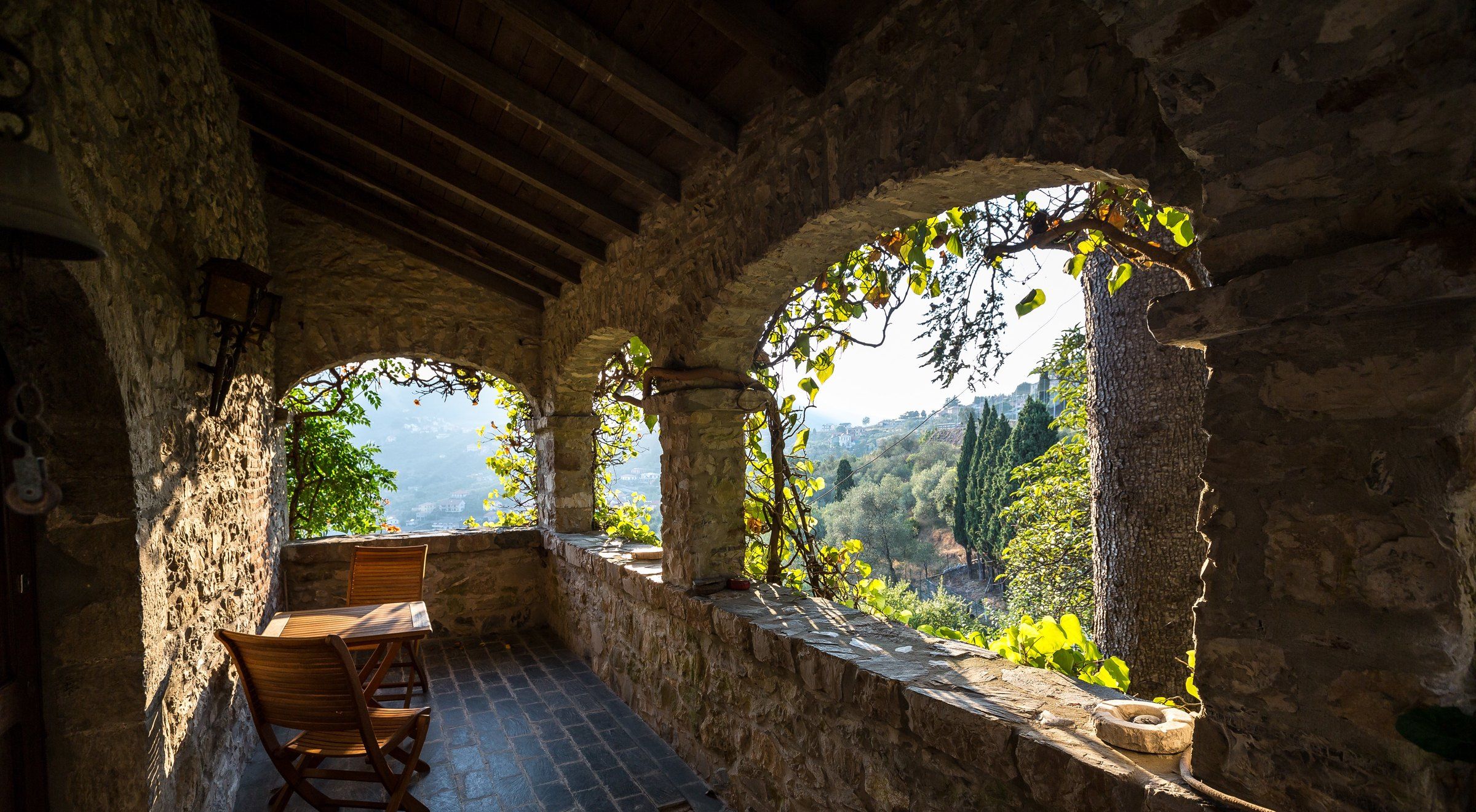 A rustic stone interior with a wooden table and chairs, stone walls, and a view of a mountainous landscape through arched openings.