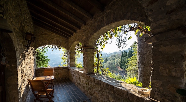 A rustic stone interior with a wooden table and chairs, stone walls, and a view of a mountainous landscape through arched openings.