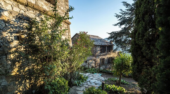 Um edifício de pedra com fachada rústica, rodeado por vegetação exuberante e um caminho de pedra que leva a uma estrutura de madeira.