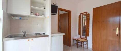 Une cuisine compacte équipée d’un évier, d’un petit réfrigérateur et d’un four à micro-ondes. On y trouve une porte en bois avec un miroir et une chaise avec un coussin.