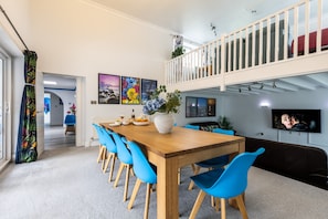 Een eethoek met een houten tafel en blauwe stoelen, een balkon met een hekwerk en een aan de muur bevestigde televisie.