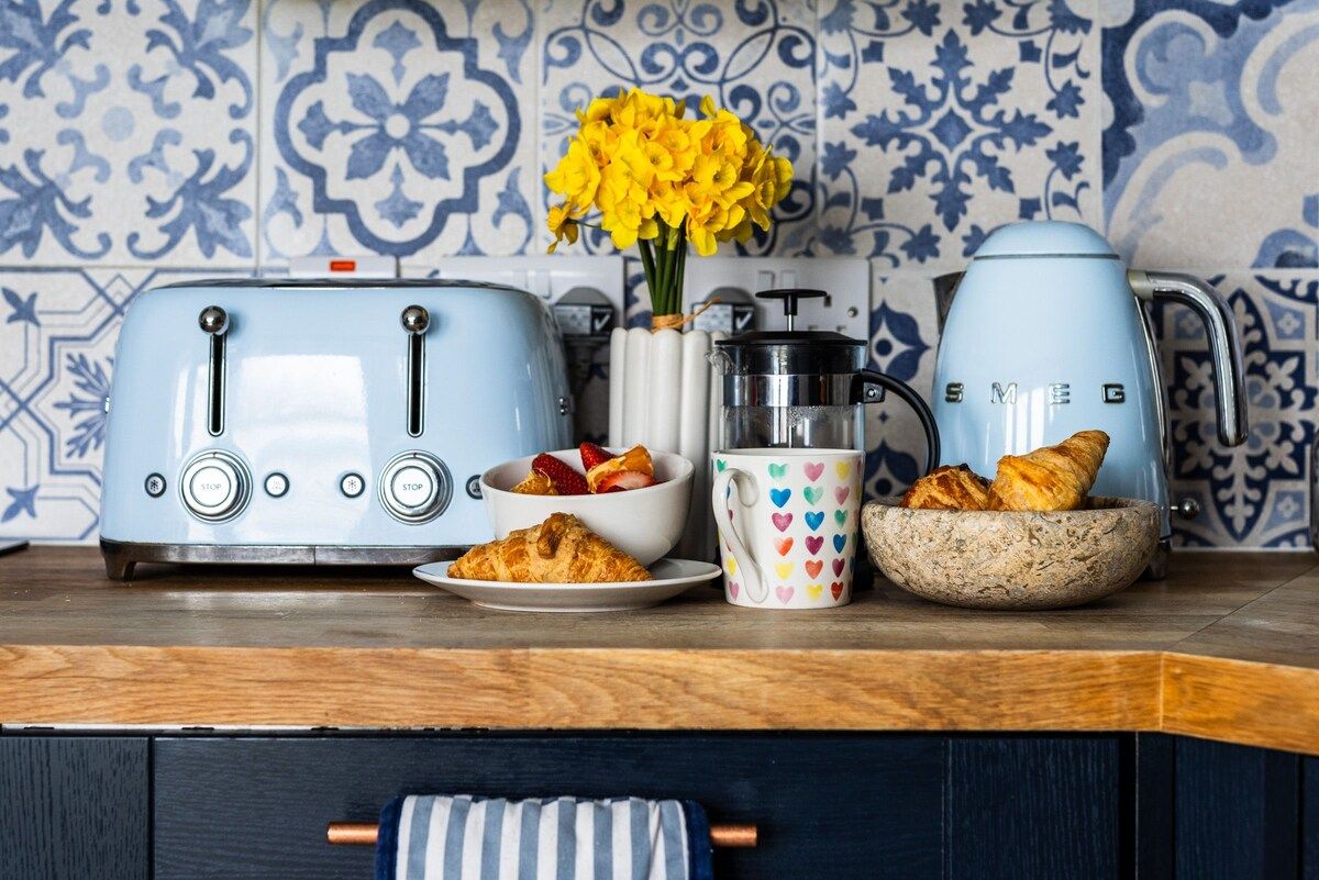 A kitchen counter with two blue toasters, a coffee maker, a mug with heart patterns, a bowl of croissants, and a vase with yellow flowers.