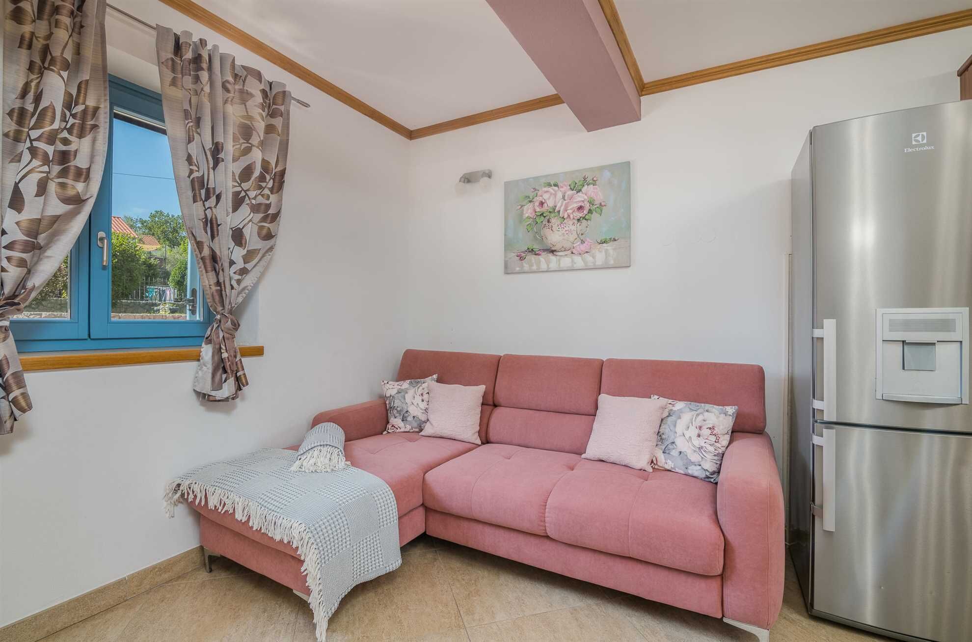 Ein gemütliches Wohnzimmer mit einem rosafarbenen Sofa, einem kleinen Tisch mit Decke, einem Kühlschrank und einem Fenster mit Vorhängen.