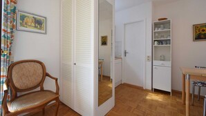 Et værelse med en træstol, et hvidt skab, et spejl, et lille bord og en hylde med køkkenudstyr.