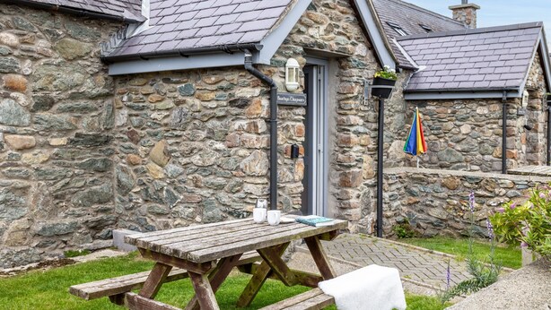 A stone building with a wooden picnic table and a door. - Bwthyn Dwynwen: Rustic Pet-Friendly Cottage in Aberffraw | Managed by Anglesey Holiday Lettings (Ty Croes)