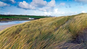 A coastal landscape with grassy dunes, a body of water, and a white building in the distance. - Bwthyn Dwynwen: Rustic Pet-Friendly Cottage in Aberffraw | Managed by Anglesey Holiday Lettings (Ty Croes)