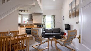 A spacious living area with a dining table, two chairs, a sofa, and a chess table. A kitchen is visible in the background with a microwave and cabinets. - Bwthyn Dwynwen: Rustic Pet-Friendly Cottage in Aberffraw | Managed by Anglesey Holiday Lettings (Ty Croes)