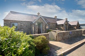 A stone building with a sloped roof, a wooden gate, and a paved driveway. - Bwthyn Dwynwen: A two bed, pet-friendly cottage in Aberffraw (Ty Croes)