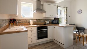 A compact kitchen with white cabinets, a built-in oven, a microwave, and a window providing natural light. - Bwthyn Dwynwen: Rustic Pet-Friendly Cottage in Aberffraw | Managed by Anglesey Holiday Lettings (Ty Croes)