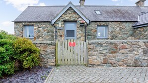 A stone building with a wooden gate, a small window, and a sign that reads "Stay with us." - Bwthyn Dwynwen: Rustic Pet-Friendly Cottage in Aberffraw | Managed by Anglesey Holiday Lettings (Ty Croes)