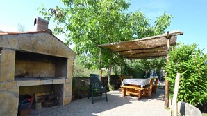 Et udendørs køkken i sten med skorsten, et overdækket siddeområde med bord og stole, og en pergola der giver skygge.