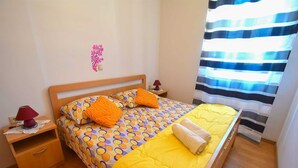 A neatly made bed with a yellow bedspread and orange pillows, a wooden nightstand with a lamp, and a striped curtain.