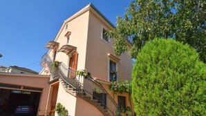A two-story building with a staircase leading to a balcony, surrounded by greenery and a parked car.
